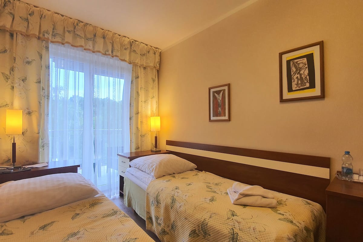 Ein Hotelzimmer mit zwei Einzelbetten, einem Nachttisch mit Lampe, einem Beistelltisch mit Wasserflasche und zwei gerahmten Bildern an der Wand.