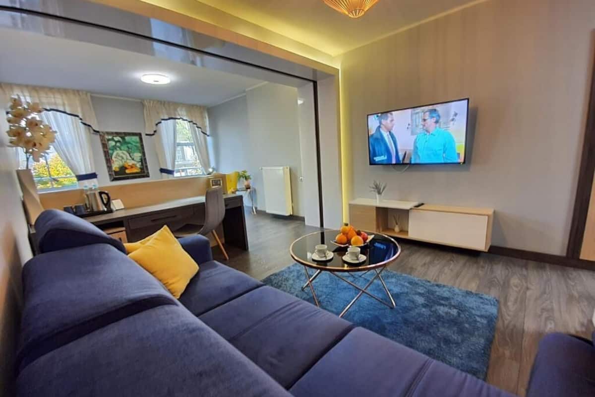 Ein modernes Wohnzimmer mit einer Couch, einem Couchtisch mit Früchten, einem Fernseher an der Wand und einem großen Spiegel, der den Raum reflektiert.