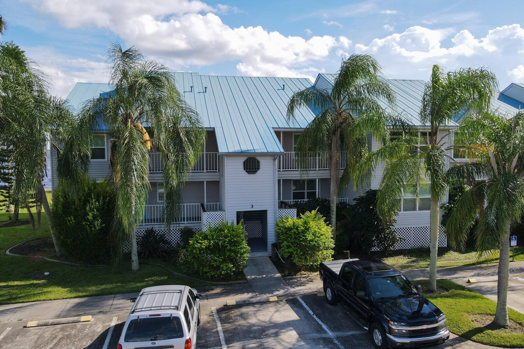 A multi-story residential building with a blue roof, surrounded by palm trees and a parking area with two vehicles.