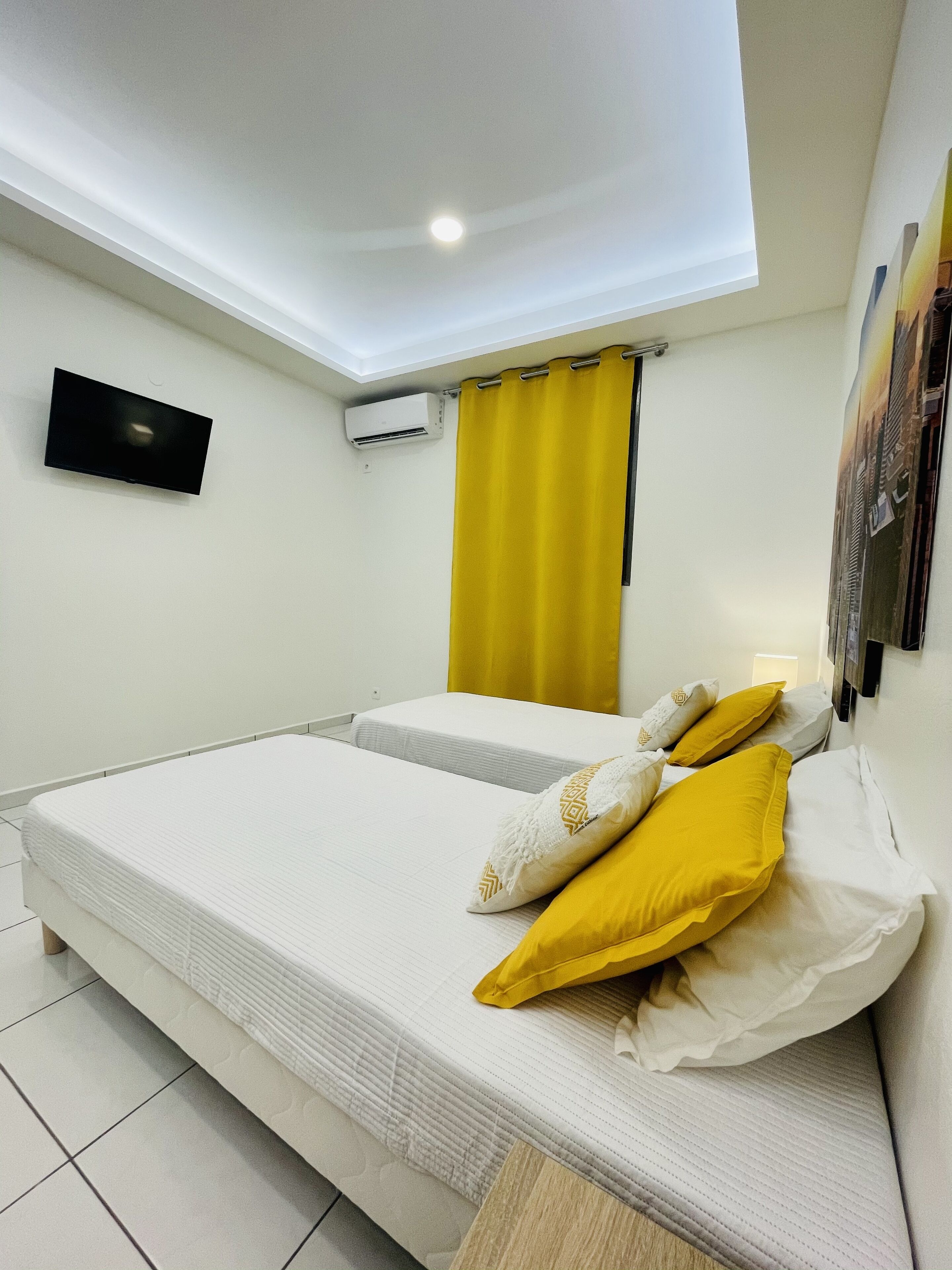 A hotel room with a bed, a flat-screen TV, an air conditioning unit, and a window with yellow curtains.