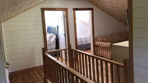 A loft bedroom with a sloped ceiling, wooden railings, a bed with a headboard, a striped sofa, and a small table. - Charming Historic Mackinaw City Cottage (Mackinaw City)