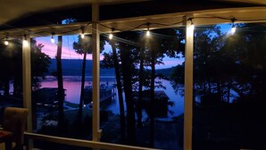 A view through a window with string lights, a wooden deck, and a body of water at dusk. - Honeycomb Waterfront Lakehouse with deep water dock (Grant)