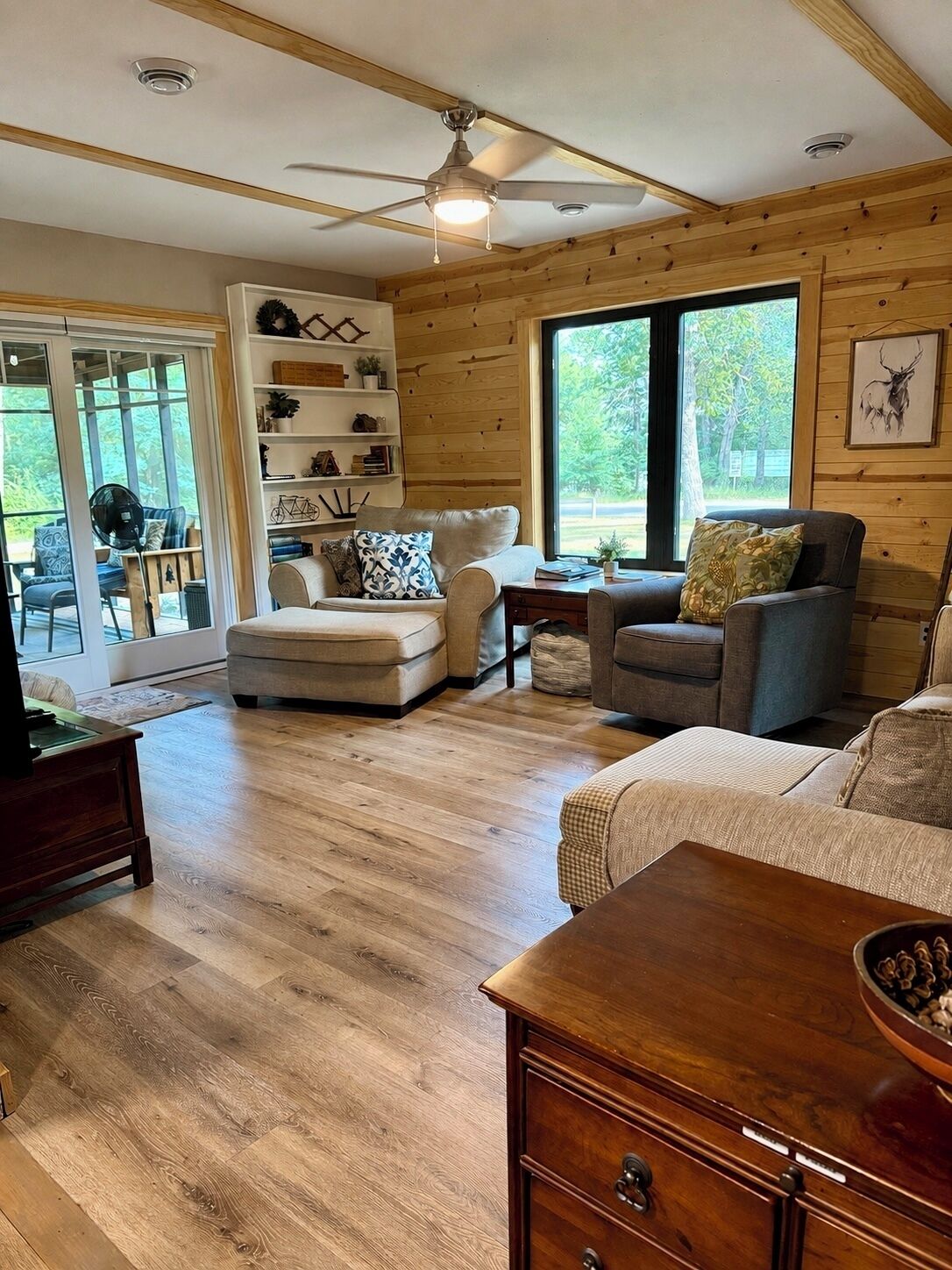 A cozy living room with a ceiling fan, wooden flooring, and a large window offering a view of the outdoors.