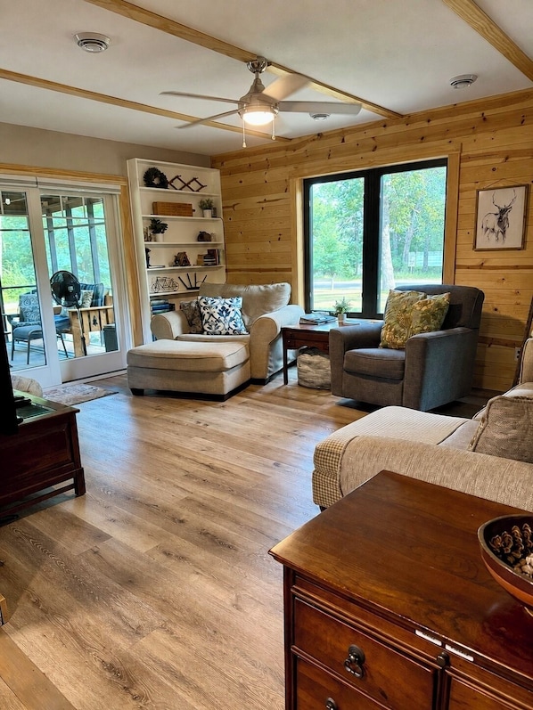 A cozy living room with a ceiling fan, wooden flooring, and a large window offering a view of the outdoors.