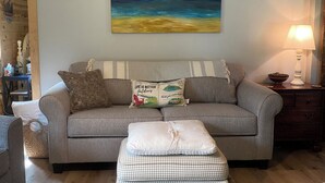 Ein Wohnzimmer mit grauem Sofa, einem karierten Hocker, einem Bild von einem Strand und einer Lampe auf einem Nachttisch aus Holz.