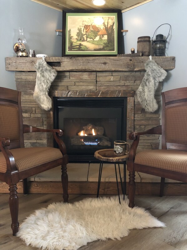 Un salon chaleureux avec une cheminée, deux fauteuils en bois, une petite table avec une tasse et un tableau encadré au-dessus du manteau de cheminée.