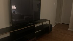 A room with a flat-screen TV on a black stand, wooden flooring, and a door in the background. - Cozy 3 bedroom house , pet friendly , yard (Portsmouth)