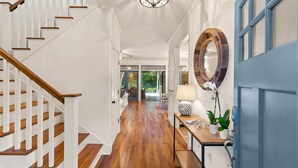 Um corredor com escada de madeira, espelho redondo, mesa com abajur e uma porta azul.