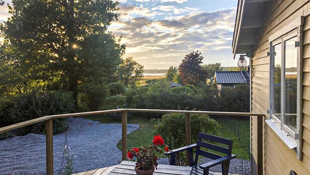 A wooden deck with a table and chairs, a potted plant with red flowers, and a view of a lake and trees in the background. - 2 bedroom cozy home in Ramdala (Ramdala)
