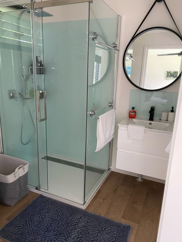 Ein modernes Badezimmer mit einer Duschkabine aus Glas, einem runden Spiegel, einem weißen Waschbecken und einer kleinen Mülleimer.