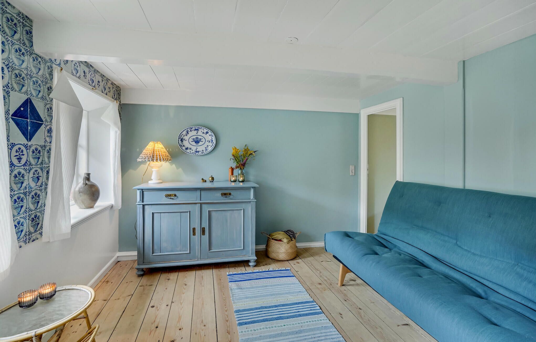 Une pièce avec un canapé bleu, une lampe blanche posée sur une commode bleue, une table en osier avec des bougies et un tapis à motifs.