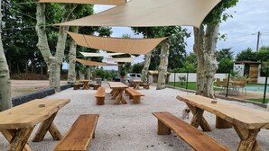 Zona de estar al aire libre con bancos de madera y mesas de picnic, protegida por grandes toldos, rodeada de árboles y un área de piscina cercada.