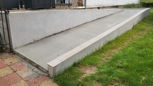 Una escalera de concreto que lleva a la entrada de un edificio, con mobiliario exterior y un  área verde cercana.