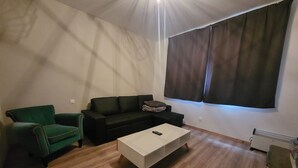 Una sala de estar con un sillón verde, un sofá negro, una mesa de centro blanca y un ventanal con cortinas oscuras.