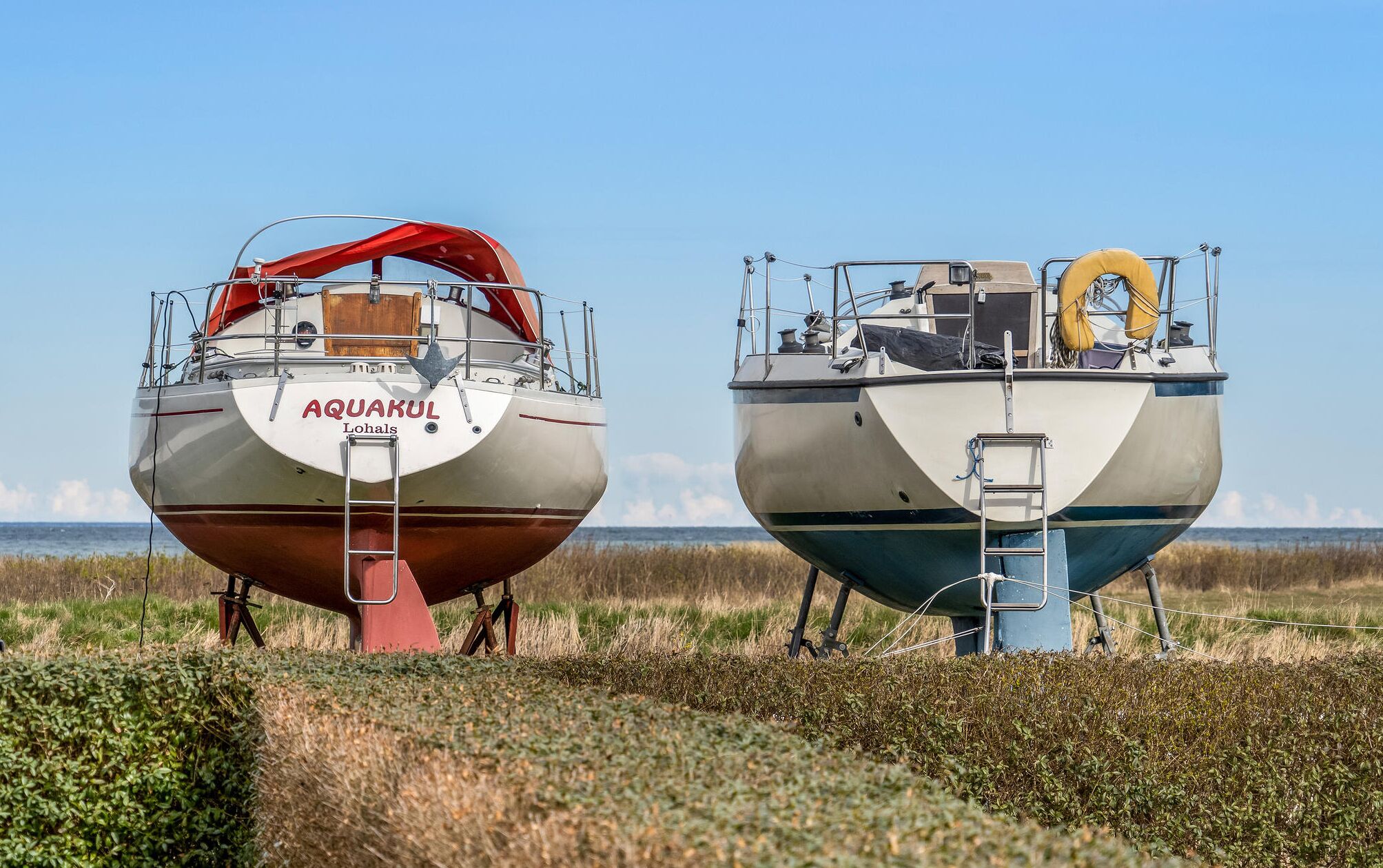 Twee stilgelegde boten met ladders en veiligheidsuitrusting, genaamd AQUAKUL Lohals, aangemeerd in een grasrijk gebied met een heldere lucht op de achtergrond.