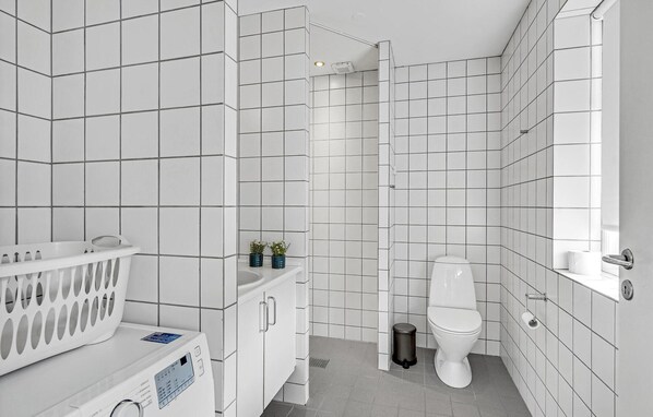 Uma casa de banho moderna com paredes e chão em azulejo branco, sanita branca, área de duche e máquina de lavar roupa.