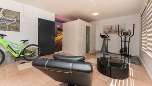 Ein Zimmer mit einem Fahrrad, einem Massagesessel, Trainingsgeräten und einer Tür mit einem malerischen Wandgemälde.