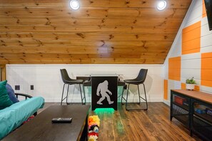 A room with wooden ceiling, a black silhouette artwork, a table, two chairs, a sofa, and a small plant on a side table.