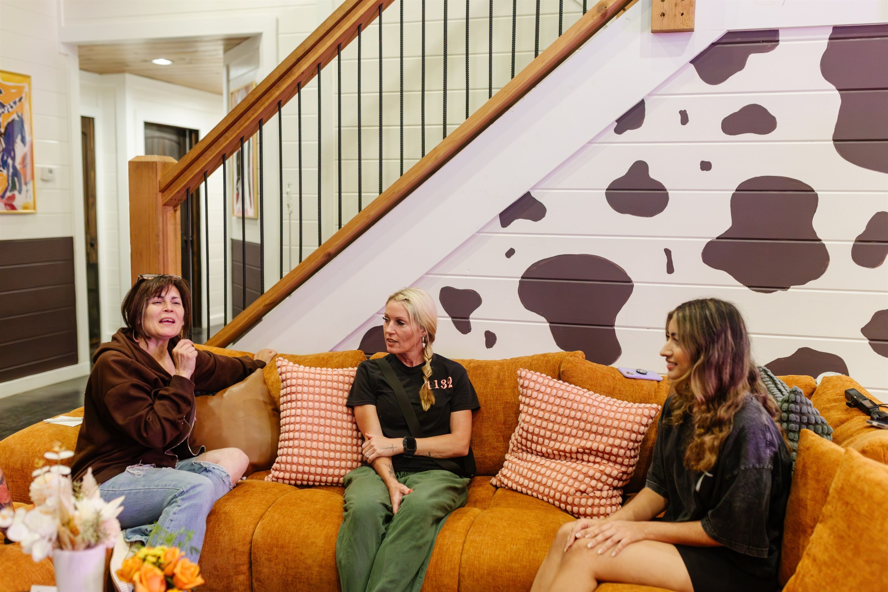 Three people sitting on an orange couch with patterned cushions in a room with a staircase and a wall with a cow print design.