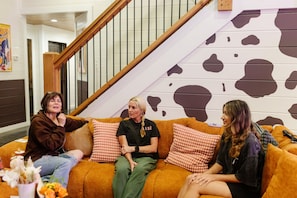 Trois personnes sont assises sur un canapé orange orné de coussins à motifs, dans une pièce qui comprend un escalier et un mur décoré d’un motif représentant des vaches.