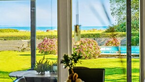 Vista de um quarto com grandes janelas para um jardim bem cuidado, com piscina, um banco e uma mesa com cadeiras.