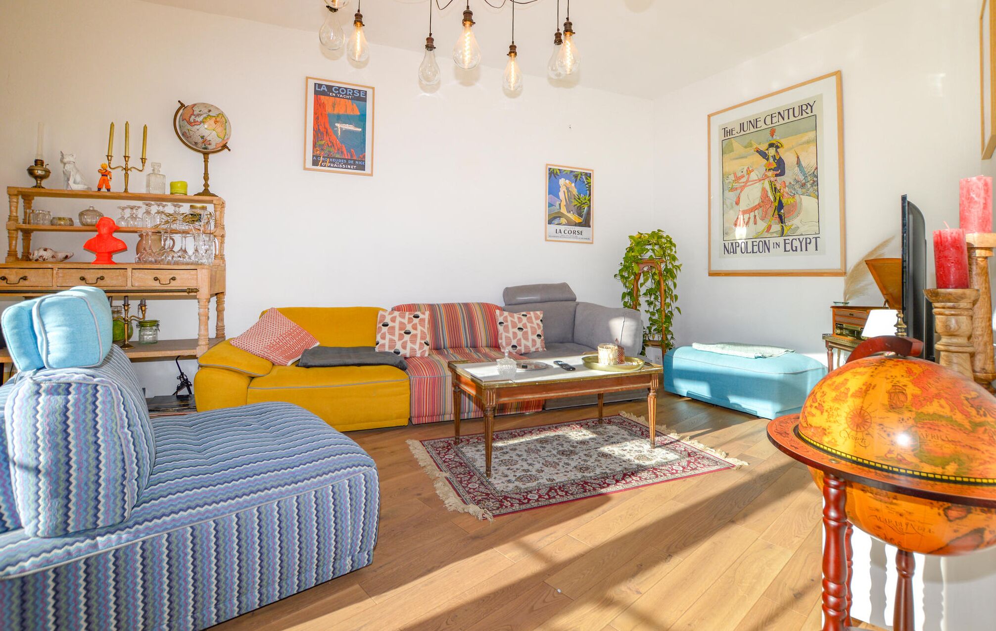Un salon chaleureux avec un canapé jaune, un fauteuil bleu et un pouf bleu. On y trouve un globe terrestre posé sur un socle en bois, une carte accrochée au mur et une affiche représentant Napoléon en Égypte.