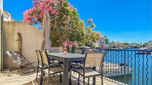 Une terrasse extérieure avec une table et des chaises, un arbre à fleurs roses et une vue sur l’eau et les bâtiments en arrière-plan.