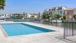 Une piscine dotée d’une terrasse en bois, entourée d’une clôture métallique et située à côté d’une marina où sont amarrés des bateaux.