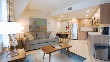 A modern living room with a sofa, coffee table, and lamp. A kitchen with wooden cabinets and appliances is visible in the background.