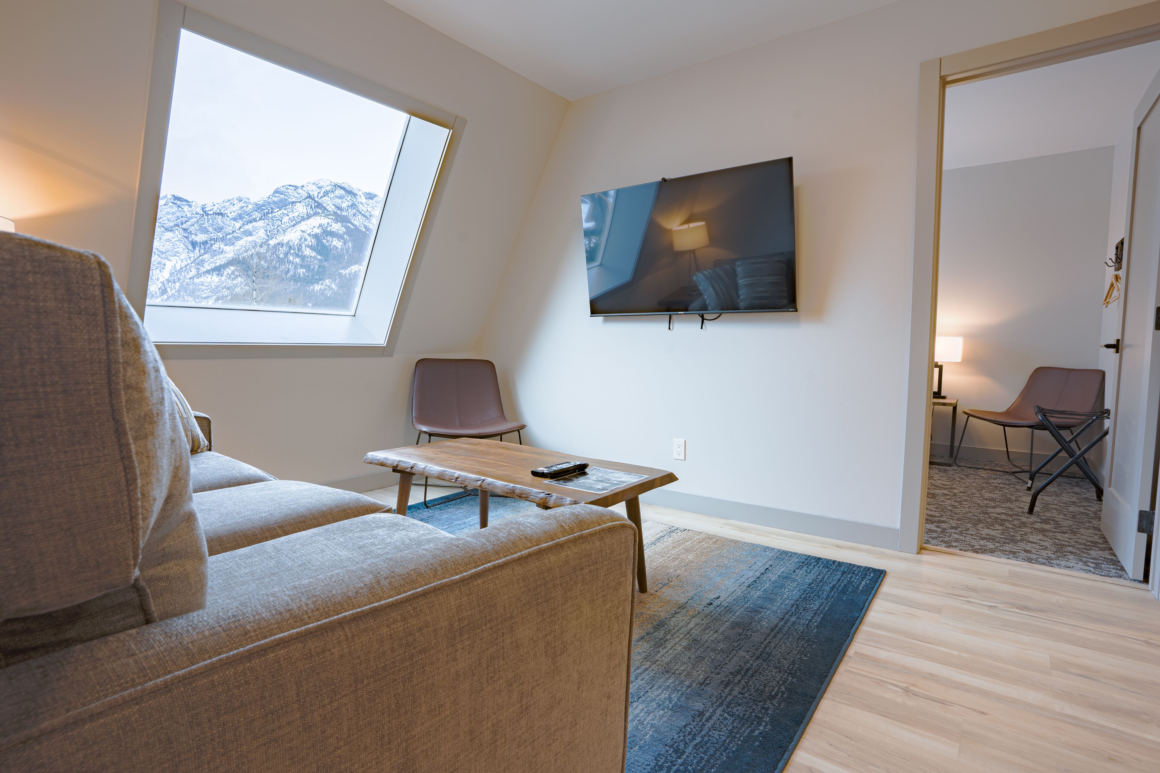 Uma sala de estar moderna com um sofá, uma mesinha e duas cadeiras. Uma TV está fixada na parede, e há uma janela com vista para montanhas cobertas de neve.