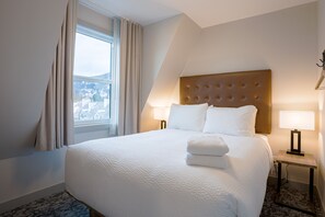 Habitación de hotel con una cama grande, una ventana con cortinas, una mesita de noche con lámpara y vistas a edificios.