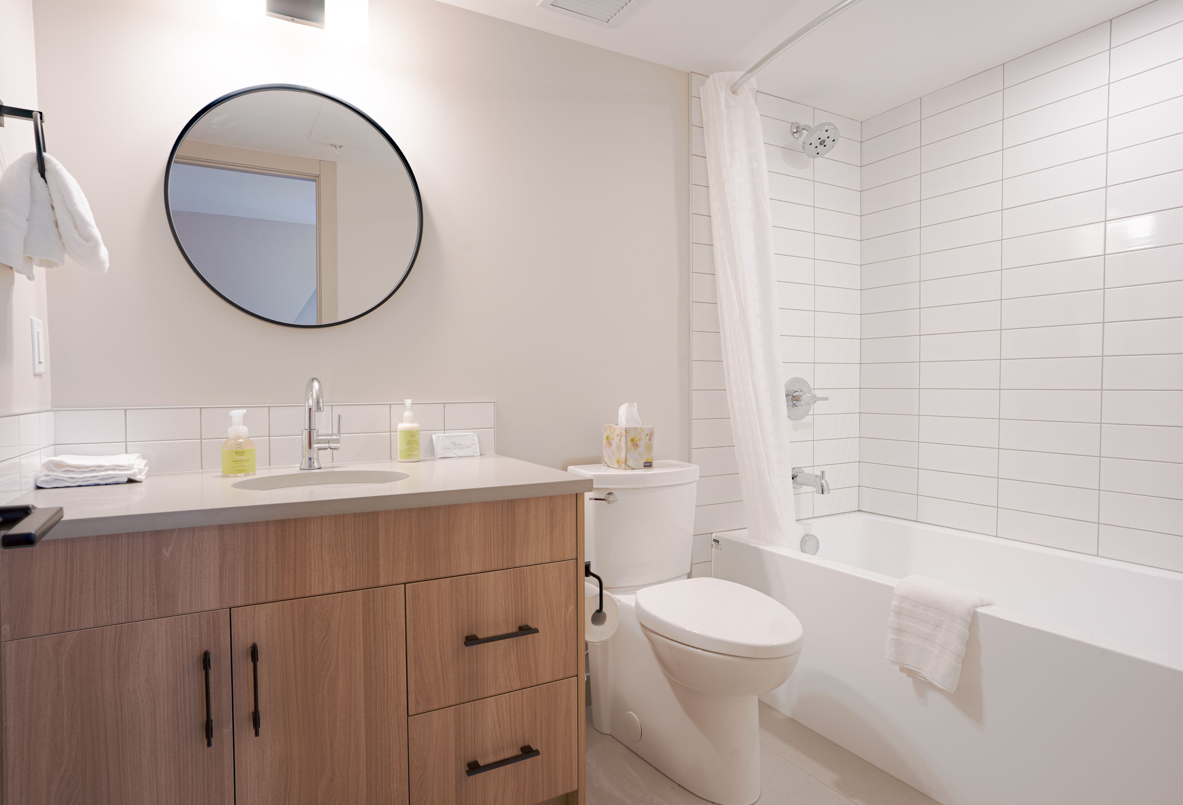 Une salle de bain moderne avec un meuble-lavabo en bois, un miroir rond, des toilettes et une baignoire dotée d’un rideau de douche.