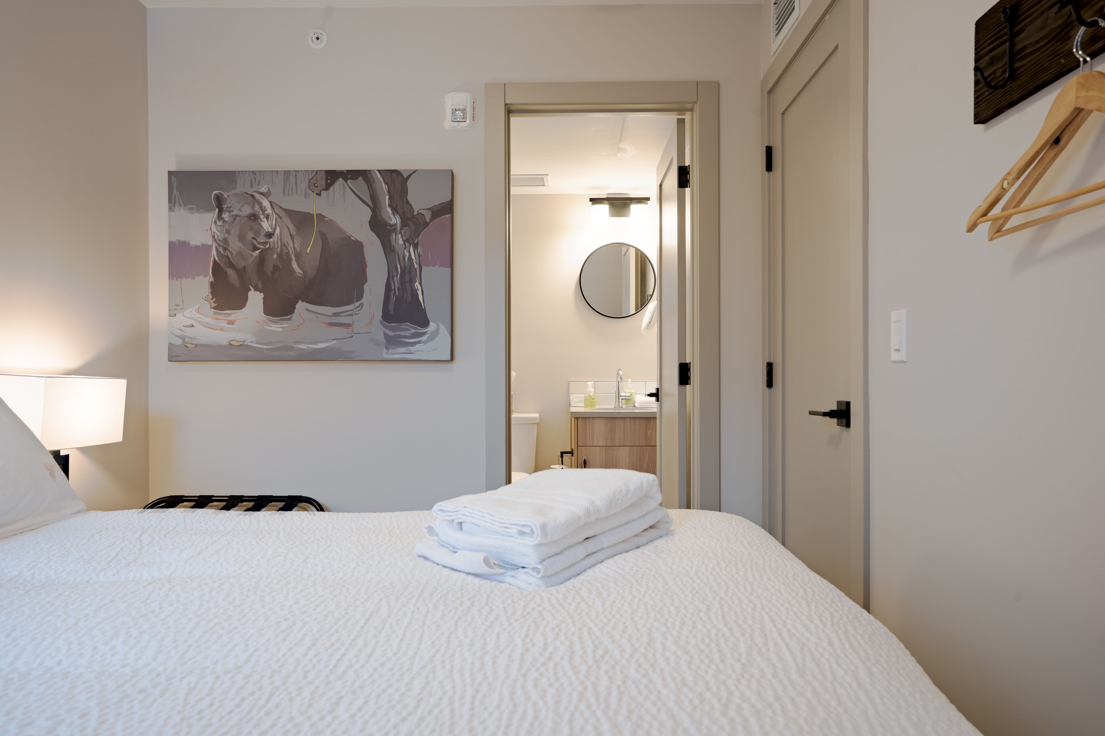 A hotel room with a bed, a painting of a bear, a round mirror, and a wooden hanger on the wall.