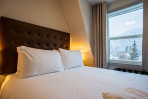 Una cama impecablemente arreglada con cabecera capitonada, una ventana con vistas a montañas nevadas y una cálida fuente de luz en la pared.