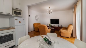 Una cocina moderna con microondas, horno y refrigerador. Hay una mesa de comedor con mantel blanco y copas de vino. Dos sillones y un sofá se encuentran en la sala de estar, que cuenta con un televisor y un reloj en la pared.