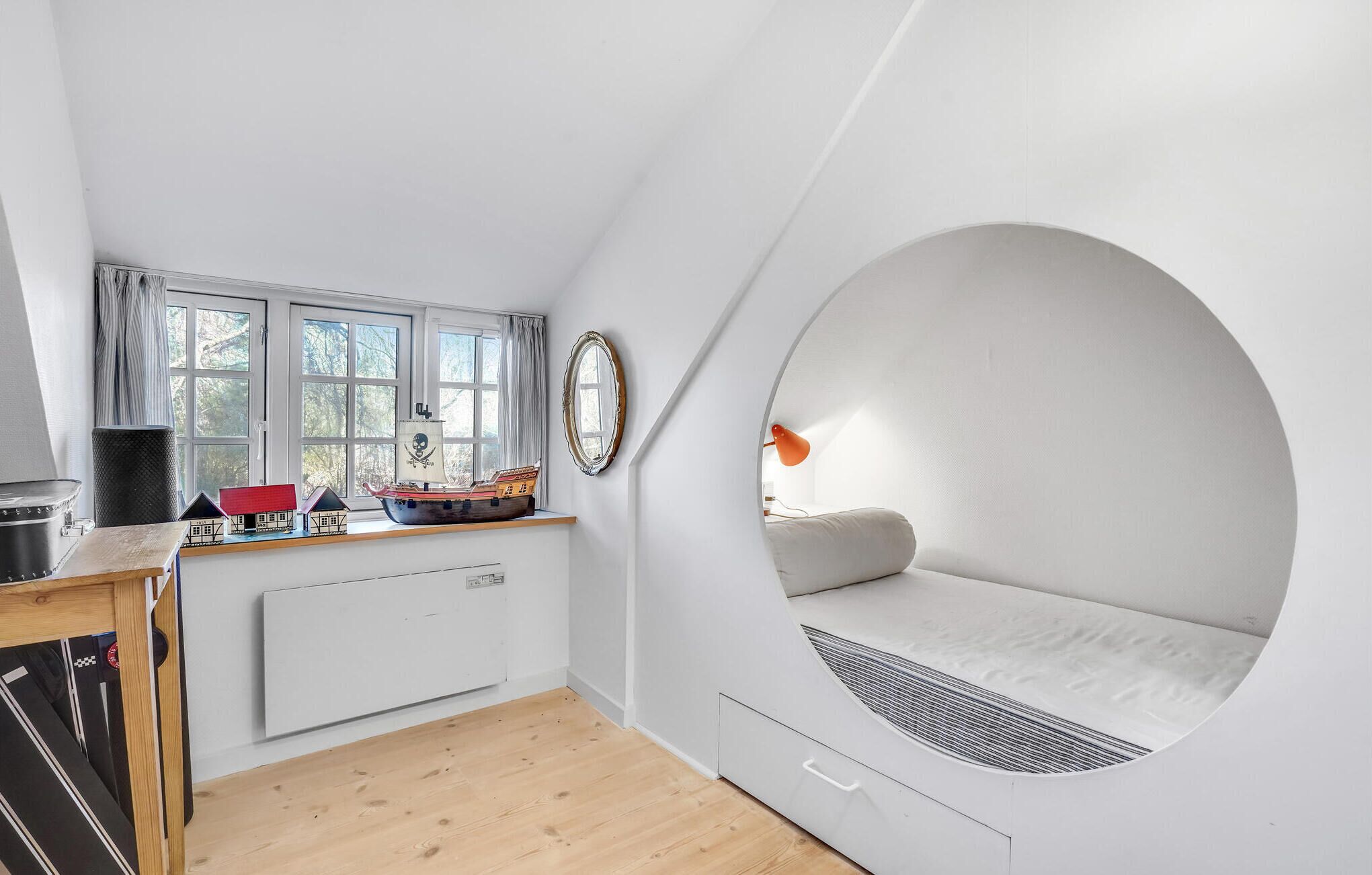 Una habitación compacta con un ventanal circular, una pequeña mesa con un modelo de barco, un radiador y una cama con cabecera.