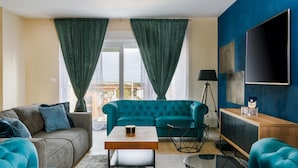 A modern living room with a teal sofa, a grey couch, and a wooden coffee table. There is a black floor lamp, a TV mounted on the wall, and a view of the outside through large windows with green curtains.
