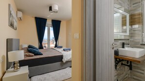 A hotel room with a bed, a bedside lamp, a nightstand, a window with curtains, and a bathroom with a sink and mirror.
