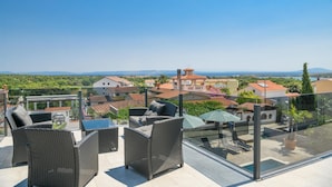 A rooftop terrace with wicker furniture, a glass balustrade, and a view of a coastal town.