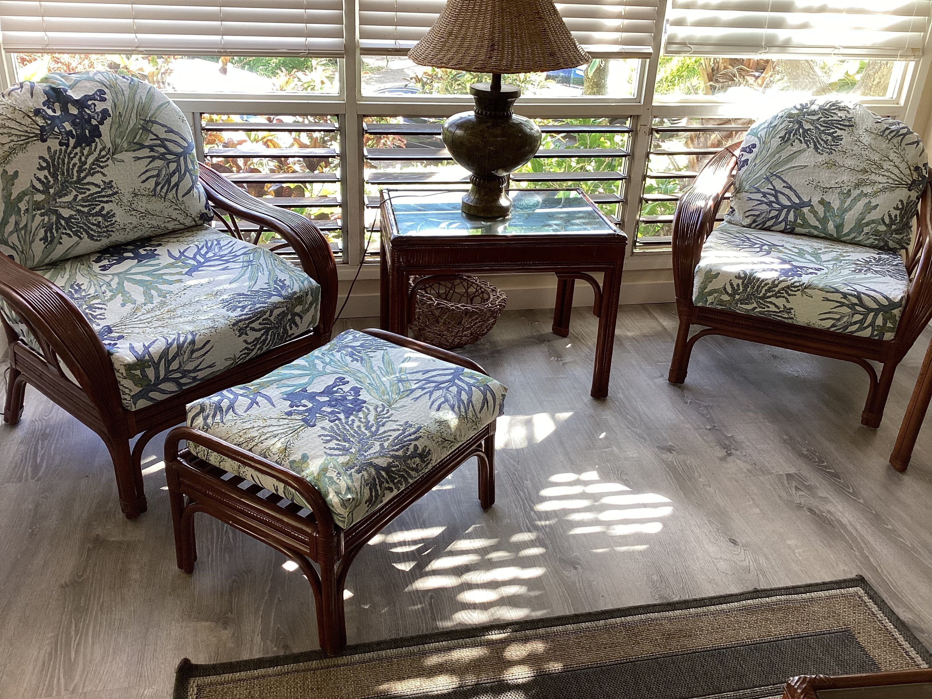A sunlit room with three upholstered chairs, a small table with a lamp, and a view through large windows.