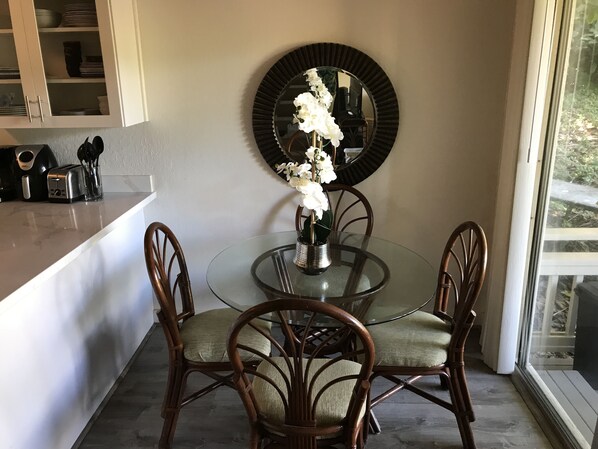 A dining area with a round glass table, four wooden chairs, a vase with white flowers, a black mirror, and a kitchen counter with a toaster and utensils.