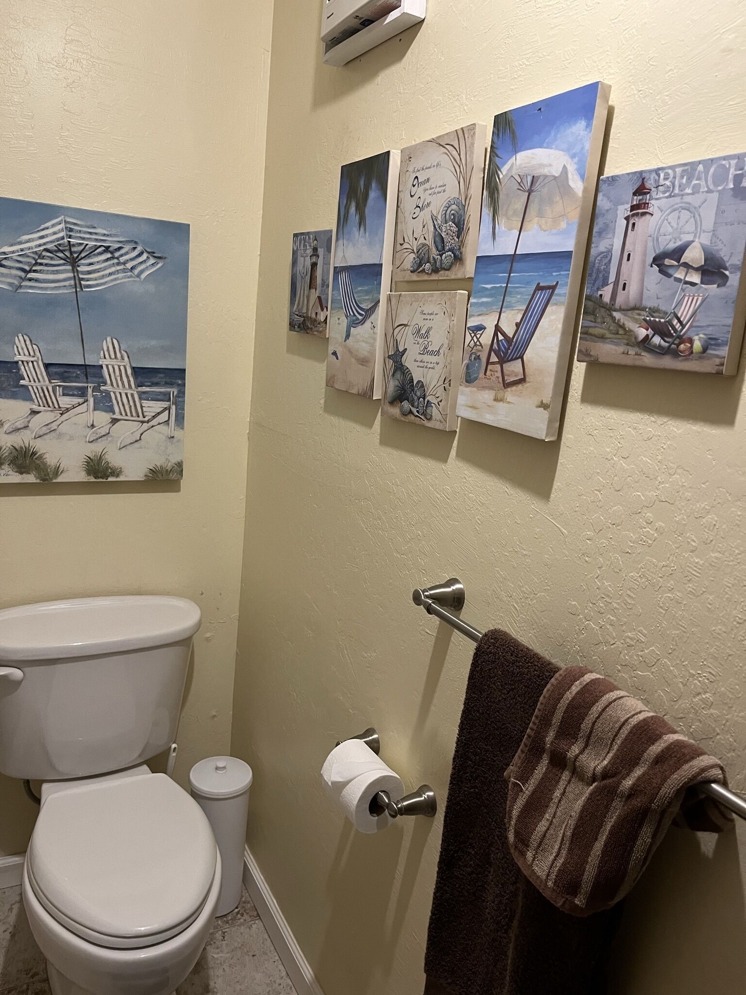 A bathroom with a toilet, a towel rack with a towel, and a series of beach-themed paintings on the wall.