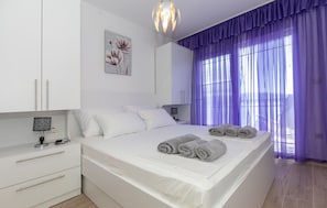 Ein modernes Schlafzimmer mit einem großen Bett, weißen Möbeln, einem dekorativen Wandbild und violetten Vorhängen.