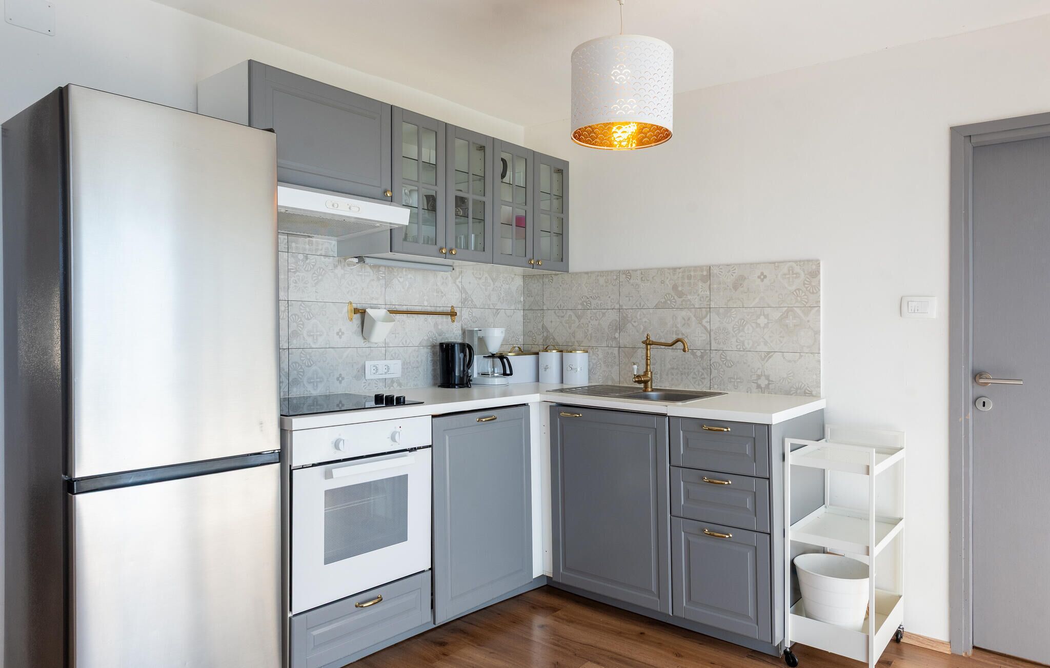 Eine moderne Küche mit grauen Schränken, einem Edelstahlkühlschrank, einem eingebauten Backofen, einer Spüle mit goldenem Wasserhahn und einer hängenden Leuchte.