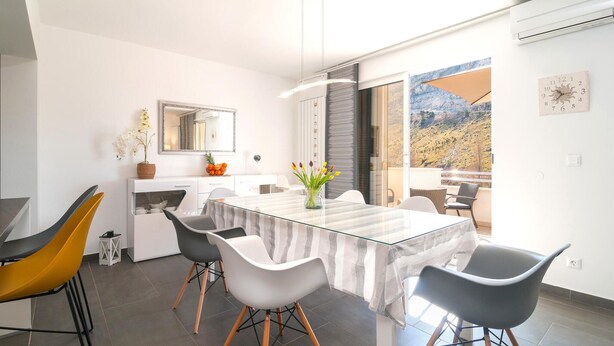 Un comedor moderno con una mesa de vidrio, sillas blancas y grises, un jarrón con flores y vistas a un paisaje montañoso a través de amplias ventanas.