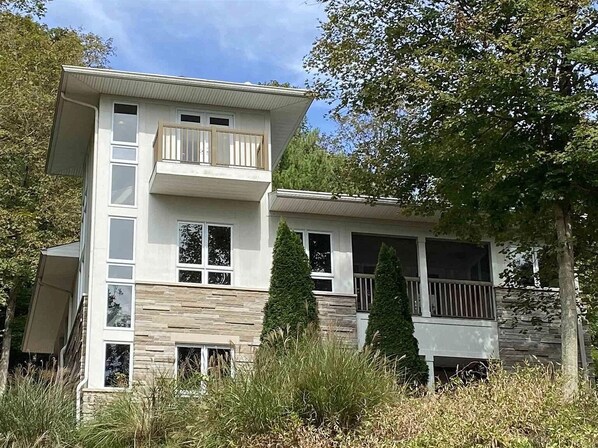A multi-story house with a balcony, stone facade, and surrounded by tall grass and trees.