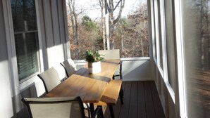 A covered patio with a wooden table and chairs, a potted plant, and a view of trees through large windows. - Beautiful new home at Kentucky Lake with community pool! Close to Murray State! (Murray)