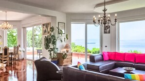 A spacious living room with a large window offering a view of the sea, a chandelier, and a modern sofa with colorful cushions. - Nice home in Kostrena (Kostrena)