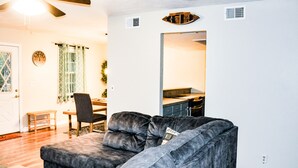 Una sala de estar con un sofá gris oscuro, una mesa de comedor de madera con sillas, un ventilador de techo y una cocina visible a través de un arco.
