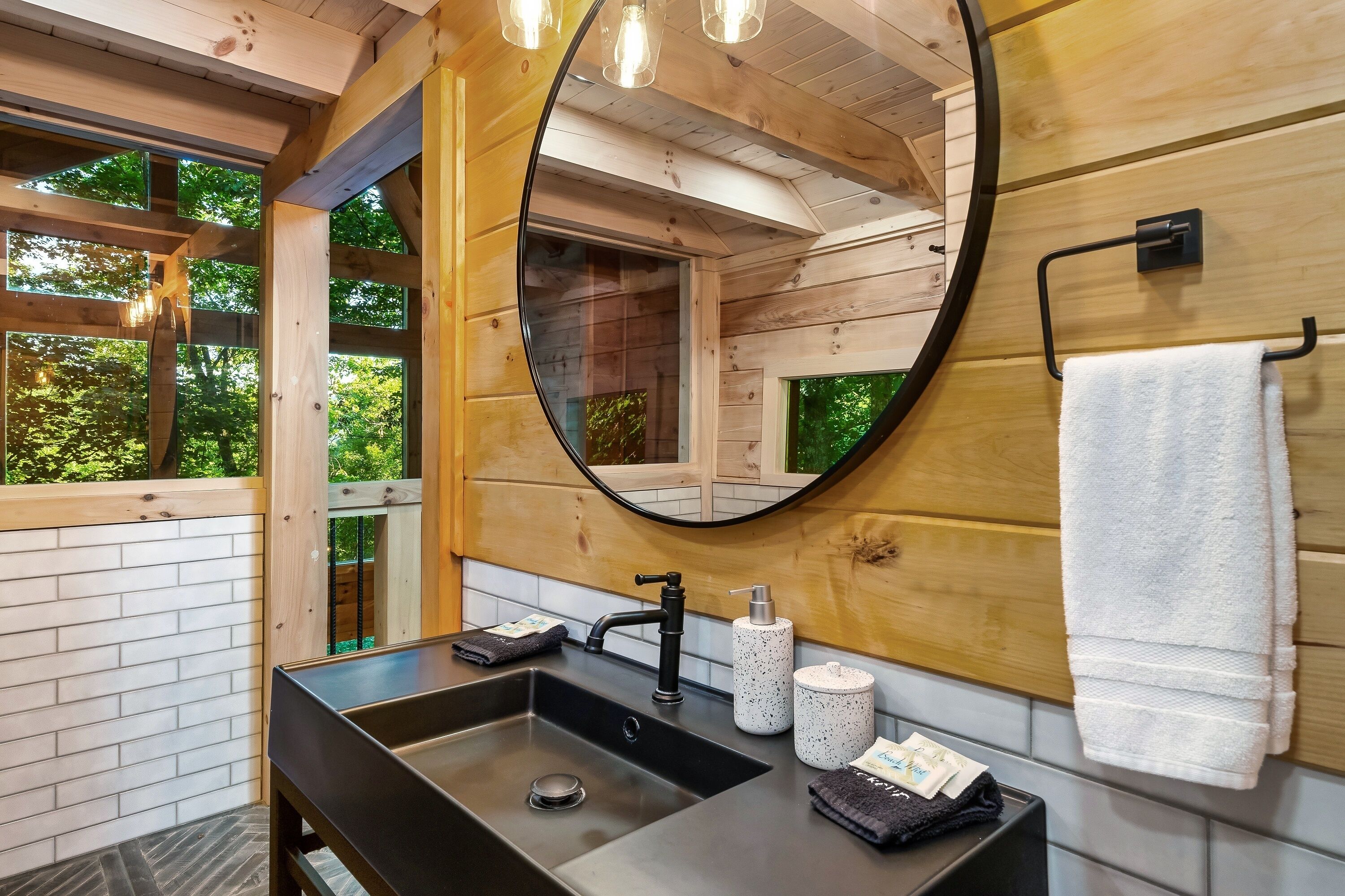 Un baño moderno con detalles de madera, un espejo redondo, un lavabo negro y un perchero para toallas.
